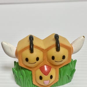 May include: A small figurine featuring three connected yellow hexagonal honeycomb cells, each with a smiling face. The top two cells have black antennae, and the bottom cell has a red mark on its forehead. The base is molded to resemble green grass.