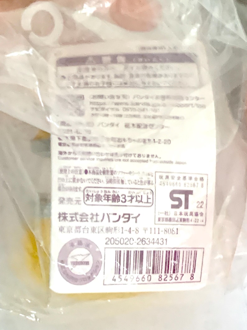 May include: A white rectangular product information tag encased in clear plastic packaging. The tag features black Japanese text, a barcode, the Bandai company name, a safety warning symbol, and the ST 22 safety certification mark for toys.