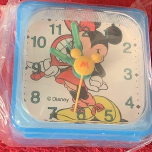 May include: A square, blue-framed alarm clock featuring a Minnie Mouse graphic. The clock face is white with black numerals and hands. The clock has a yellow Mickey Mouse head on the center. The clock is in a plastic wrapper. The Disney logo is visible.