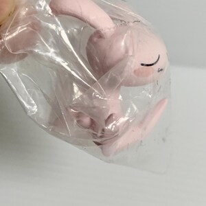 Puede incluir: Una peque&ntilde;a figura de color rosa claro de una criatura con orejas largas y expresi&oacute;n de ojos cerrados, envuelta en una bolsa de pl&aacute;stico transparente. La figura presenta un acabado mate liso con l&iacute;neas negras simples para los ojos y una boca peque&ntilde;a, en una posici&oacute;n sentada o encogida.