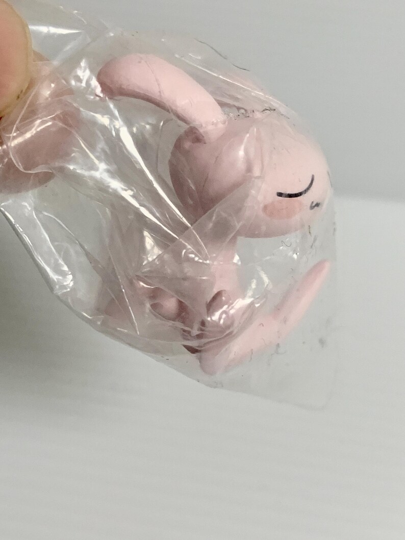 Puede incluir: Una peque&ntilde;a figura de color rosa claro de una criatura con orejas largas y expresi&oacute;n de ojos cerrados, envuelta en una bolsa de pl&aacute;stico transparente. La figura presenta un acabado mate liso con l&iacute;neas negras simples para los ojos y una boca peque&ntilde;a, en una posici&oacute;n sentada o encogida.