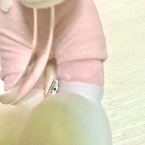 May include: A plush toy featuring a soft pink fabric body with white accents. The toy has a small tag attached near the leg area that displays a circular Disney Mickey Mouse icon. A dark green tag with the text LIMITED is visible at the top.