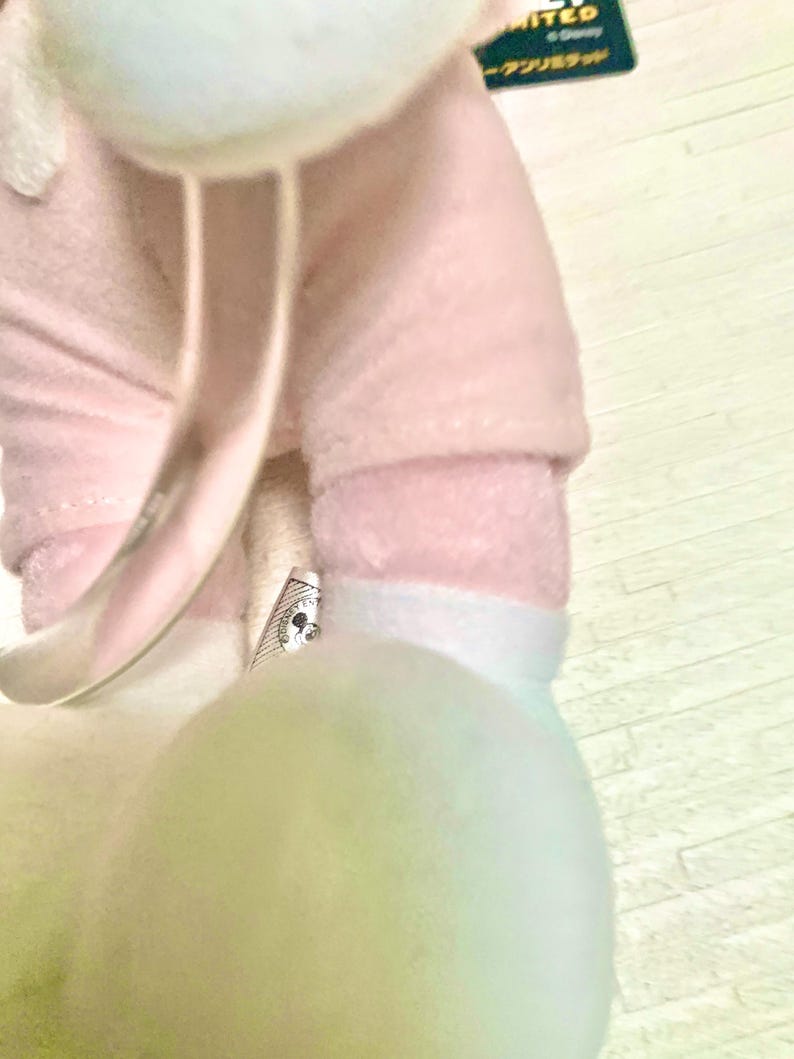 May include: A plush toy featuring a soft pink fabric body with white accents. The toy has a small tag attached near the leg area that displays a circular Disney Mickey Mouse icon. A dark green tag with the text LIMITED is visible at the top.