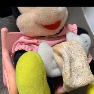 May include: A vintage Mickey Mouse plush toy, featuring a pink dress, black ears, and yellow feet. The plush is seated on a pink chair, with white gloves and a red smile. The toy's fur is a light beige color.