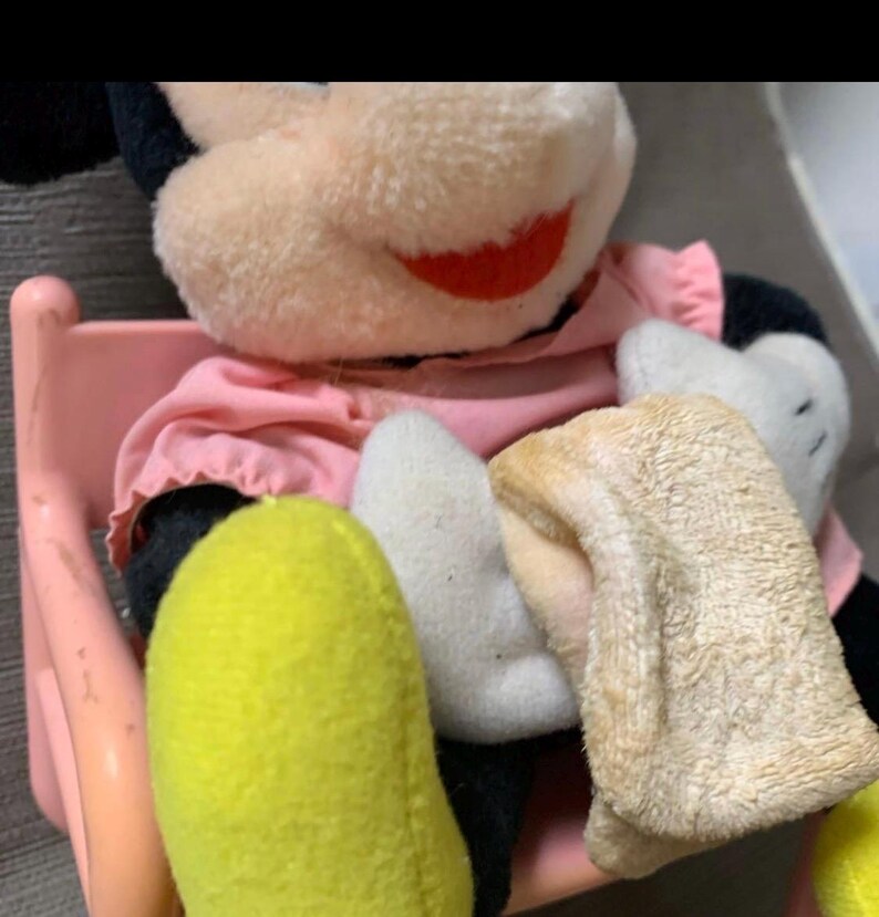 May include: A vintage Mickey Mouse plush toy, featuring a pink dress, black ears, and yellow feet. The plush is seated on a pink chair, with white gloves and a red smile. The toy's fur is a light beige color.