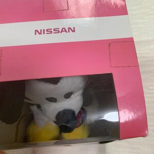 Puede incluir: Un peluche en una caja de pl&aacute;stico transparente. El juguete es blanco con manchas negras y una nariz negra. Tiene pies amarillos y una bufanda colorida. La caja es rosa con la palabra "NISSAN" impresa.
