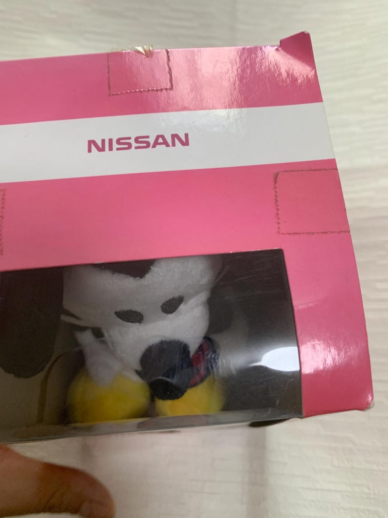 Puede incluir: Un peluche en una caja de pl&aacute;stico transparente. El juguete es blanco con manchas negras y una nariz negra. Tiene pies amarillos y una bufanda colorida. La caja es rosa con la palabra "NISSAN" impresa.