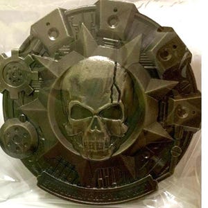 May include: A detailed, metallic-looking emblem with a central skull design. The emblem features a cracked skull, surrounded by starbursts, gears, and geometric shapes. The overall color is a muted, dark green.