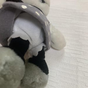 Op de afbeelding: Een zacht pandabeerspeelgoed met zwart-witte vacht, gekleed in een grijze jurk met witte stippen. De poten van de beer zijn grijs en zwart. Een schattig en knuffelbaar speelgoed voor kinderen.