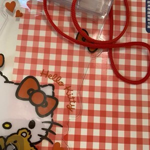 Pode incluir: Cord&atilde;o Hello Kitty de pl&aacute;stico transparente com um cord&atilde;o vermelho e um porta-identifica&ccedil;&otilde;es transparente. O cord&atilde;o apresenta um design Hello Kitty com um la&ccedil;o vermelho e um ursinho de pel&uacute;cia. O fundo &eacute; um padr&atilde;o xadrez vermelho e branco.