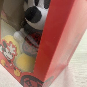 Puede incluir: Un peluche de Mickey Mouse en una caja roja con una ventana transparente. Mickey Mouse es blanco, negro y rojo. La caja tiene un gr&aacute;fico de Mickey Mouse y un patr&oacute;n de tablero de ajedrez negro y rojo en la parte inferior.