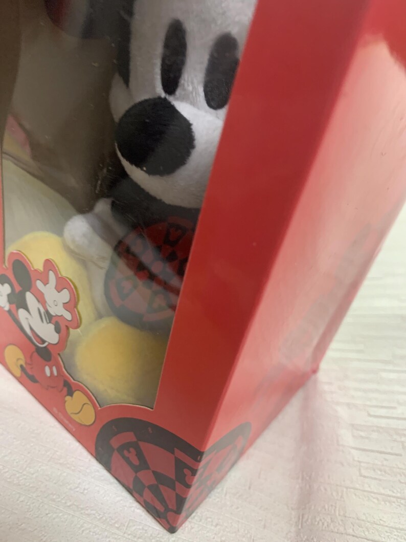 Puede incluir: Un peluche de Mickey Mouse en una caja roja con una ventana transparente. Mickey Mouse es blanco, negro y rojo. La caja tiene un gr&aacute;fico de Mickey Mouse y un patr&oacute;n de tablero de ajedrez negro y rojo en la parte inferior.