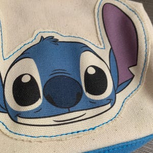 以下が含まれることがあります： 大きな黒い目と紫色の耳を持つ、青と白の漫画のキャラクターの顔が特徴のキャンバスポーチ。ポーチには青いジッパーとトリムがあり、上部には青いストラップが見えます。