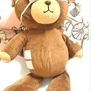 May include: A brown teddy bear toy with a beige muzzle, wearing a white eye patch and bandages. The bear has a stitched black mouth and nose. The bear's arms and legs are outstretched, and it appears to be a stuffed animal.