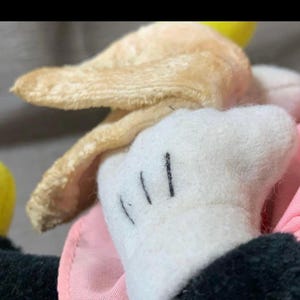 May include: Close-up of a vintage plush toy featuring a white glove with three black lines, a tan ear, and a pink fabric. The toy's black sleeve is visible.