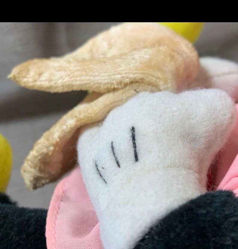 May include: Close-up of a vintage plush toy featuring a white glove with three black lines, a tan ear, and a pink fabric. The toy's black sleeve is visible.