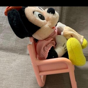 May include: A vintage Mickey Mouse plush toy wearing a pink dress and a red hat, seated on a small pink plastic chair. The plush has black ears, a white face, and yellow feet. The chair is a light pink color.