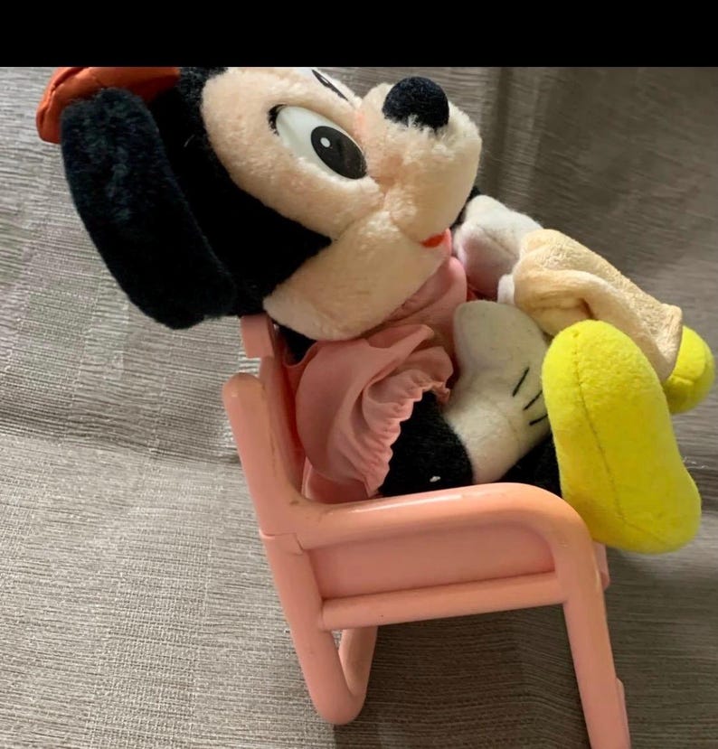 May include: A vintage Mickey Mouse plush toy wearing a pink dress and a red hat, seated on a small pink plastic chair. The plush has black ears, a white face, and yellow feet. The chair is a light pink color.
