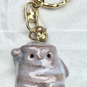 Puede incluir: Un llavero con una pequeña figura de cerámica esmaltada de forma redondeada y abstracta en tonos marrón, gris y blanco. La figura tiene dos pequeñas hendiduras oscuras para los ojos y está unida a una anilla y un cierre de mosquetón de metal dorado.