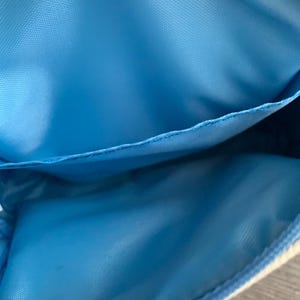 以下が含まれることがあります： 小さな水色のポーチの内側のクローズアップ。ポーチには水色の裏地と目に見える縫い目があります。素材は滑らかな合成繊維のようです。ポーチは開いており、内部が明らかになっています。