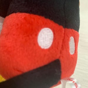 Puede incluir: Primer plano de un peluche con un cuerpo rojo, dos c&iacute;rculos blancos y una parte inferior negra. La parte superior del juguete es negra. Se adjunta una cinta blanca con texto rojo.