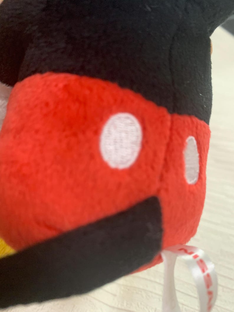 Puede incluir: Primer plano de un peluche con un cuerpo rojo, dos c&iacute;rculos blancos y una parte inferior negra. La parte superior del juguete es negra. Se adjunta una cinta blanca con texto rojo.