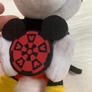 Puede incluir: Un peluche de Mickey Mouse con orejas negras, cuerpo blanco y negro, guantes y zapatos amarillos. El juguete sostiene un emblema circular rojo y negro con siluetas de Mickey Mouse.