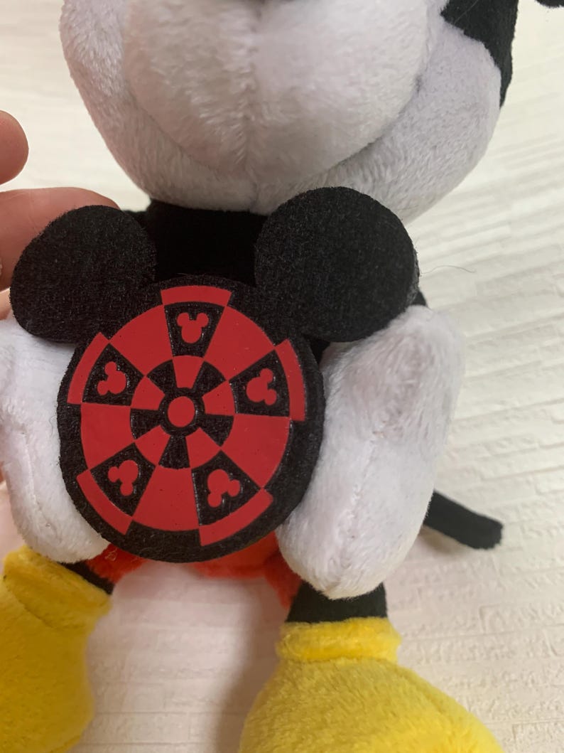 Puede incluir: Un peluche de Mickey Mouse con orejas negras, cuerpo blanco y negro, guantes y zapatos amarillos. El juguete sostiene un emblema circular rojo y negro con siluetas de Mickey Mouse.