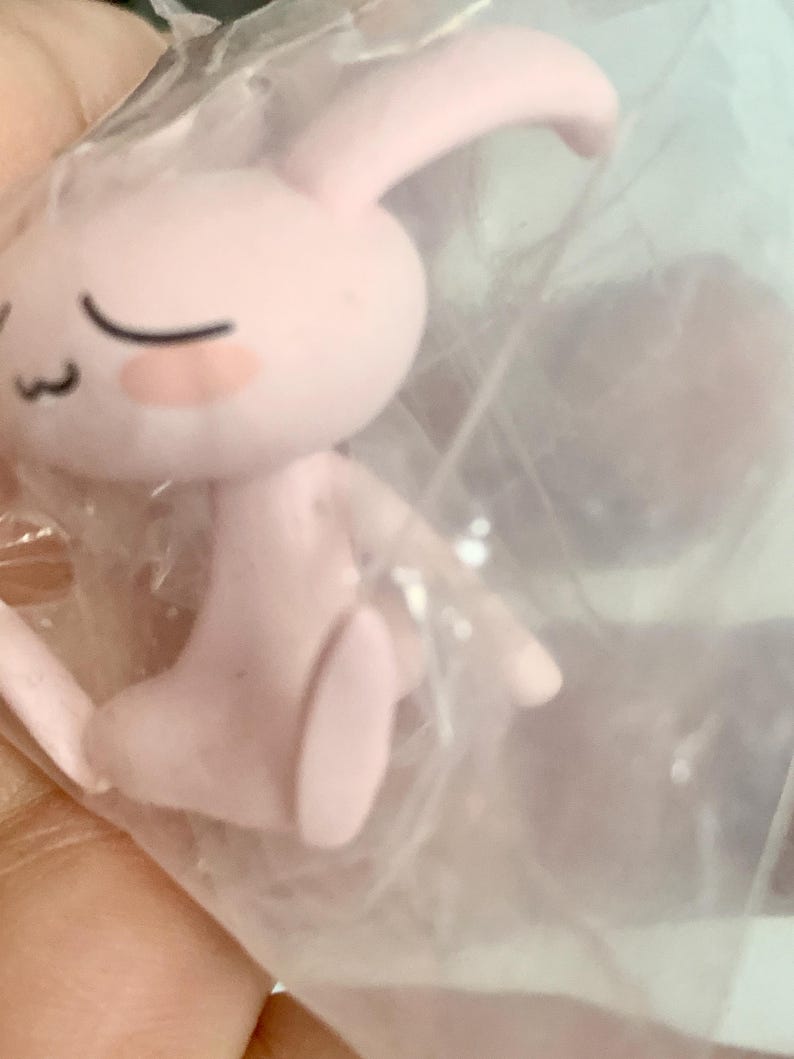 Puede incluir: Una peque&ntilde;a figura de color rosa claro de una criatura parecida a un conejo con orejas largas, ojos cerrados y una boca sencilla, envuelta en una bolsa de pl&aacute;stico transparente. El personaje presenta rasgos faciales suaves y redondeados.