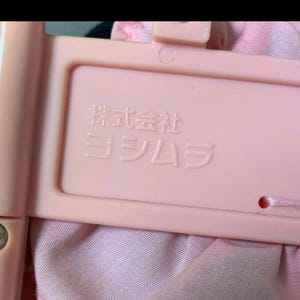 May include: Close-up of a light pink plastic frame with Japanese text. The frame is rectangular with rounded corners and a small hole. The text is embossed in white. The frame is attached to a pink fabric.