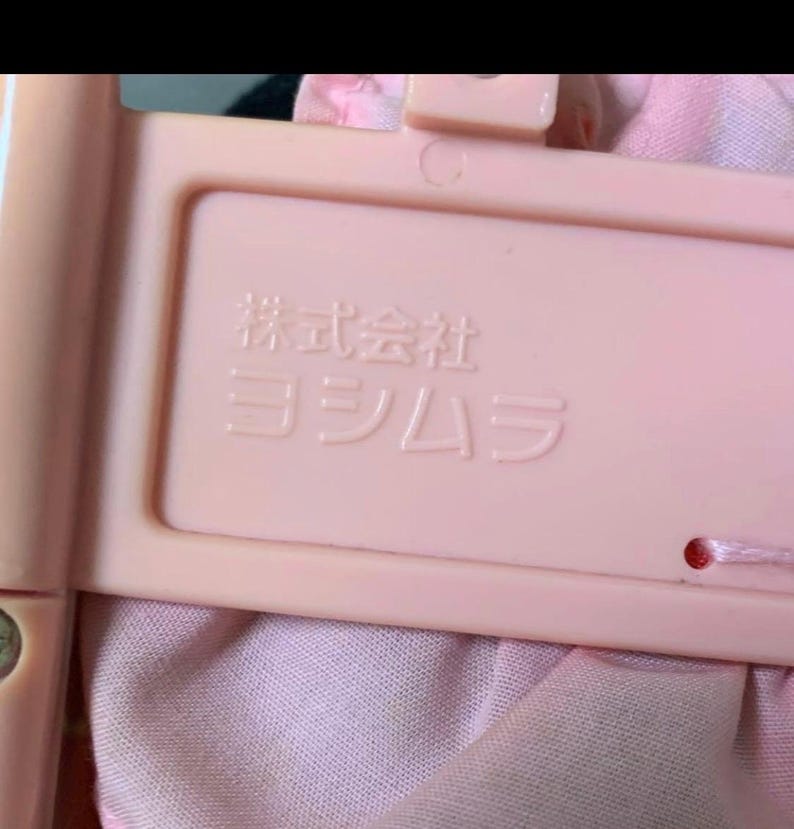 May include: Close-up of a light pink plastic frame with Japanese text. The frame is rectangular with rounded corners and a small hole. The text is embossed in white. The frame is attached to a pink fabric.