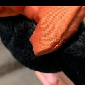 May include: Close-up of a black plush toy with an orange satin bow. The bow is tied in a simple knot and has a textured appearance. The plush toy has a soft, fuzzy texture.