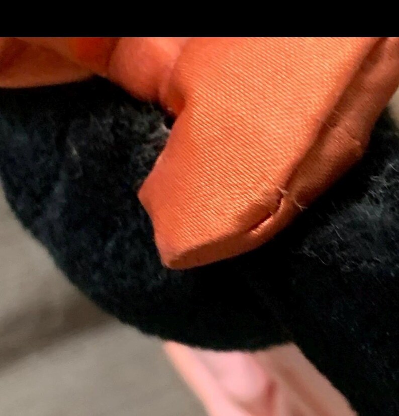 May include: Close-up of a black plush toy with an orange satin bow. The bow is tied in a simple knot and has a textured appearance. The plush toy has a soft, fuzzy texture.