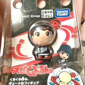 Puede incluir: Figura Jung Kook Choro Q empaquetada de la serie MIC Drop. La figura está en una caja de plástico transparente y presenta un personaje de estilo caricatura con una camisa a rayas blancas y negras y zapatos rojos. El embalaje incluye texto japonés y el logotipo de Takara Tomy.
