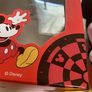 Puede incluir: Caja roja con gr&aacute;ficos de Mickey Mouse y un dise&ntilde;o de diana. La caja tiene una ventana transparente y el logotipo de Disney. La diana tiene secciones negras y rojas con la silueta de Mickey Mouse.