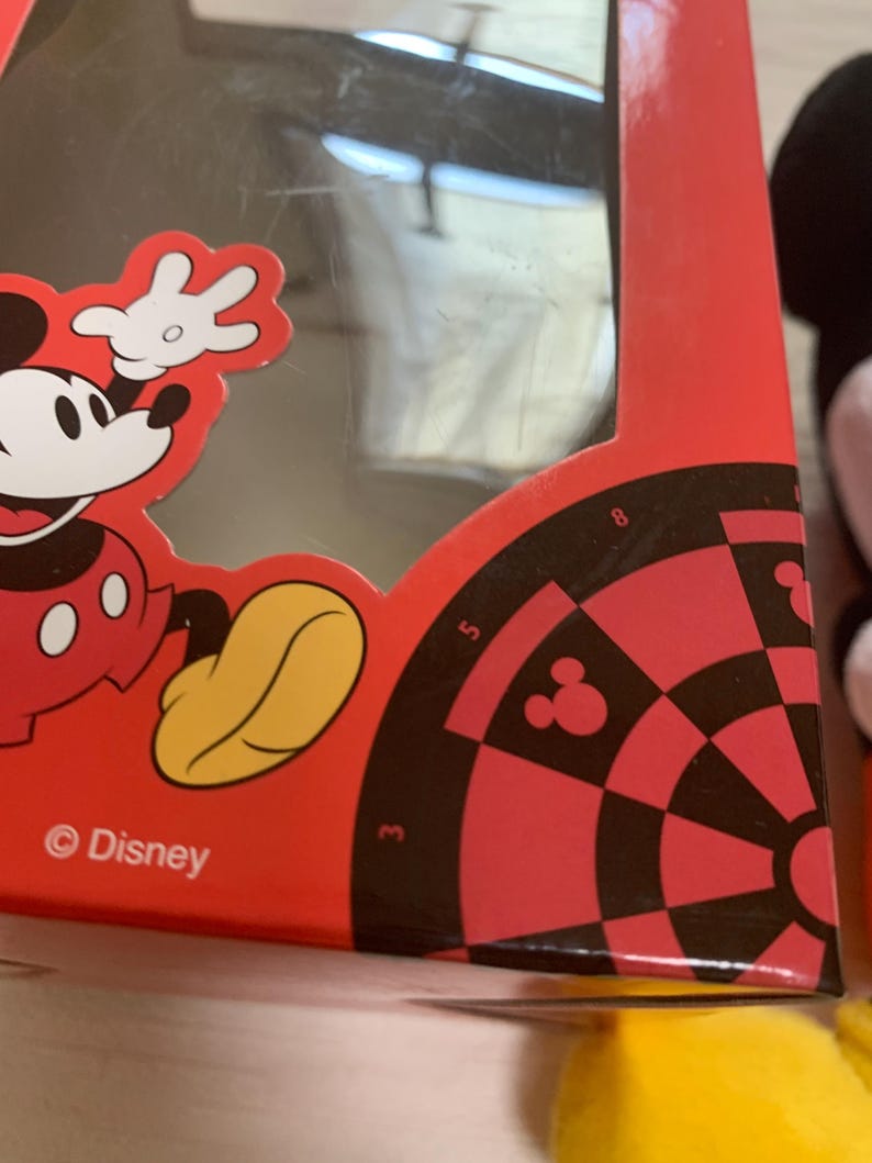 Puede incluir: Caja roja con gr&aacute;ficos de Mickey Mouse y un dise&ntilde;o de diana. La caja tiene una ventana transparente y el logotipo de Disney. La diana tiene secciones negras y rojas con la silueta de Mickey Mouse.