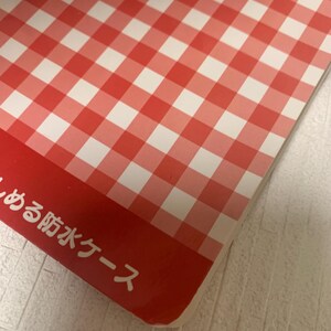 Pode incluir: Um padr&atilde;o xadrez vermelho e branco cobre a maior parte da imagem, com uma borda vermelha na parte inferior. O texto japon&ecirc;s &eacute; vis&iacute;vel na borda vermelha. O padr&atilde;o &eacute; um design xadrez cl&aacute;ssico, frequentemente usado em piqueniques.
