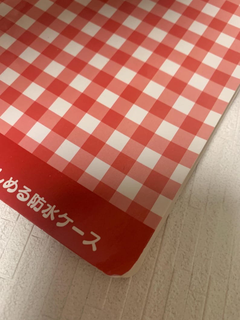 Pode incluir: Um padr&atilde;o xadrez vermelho e branco cobre a maior parte da imagem, com uma borda vermelha na parte inferior. O texto japon&ecirc;s &eacute; vis&iacute;vel na borda vermelha. O padr&atilde;o &eacute; um design xadrez cl&aacute;ssico, frequentemente usado em piqueniques.