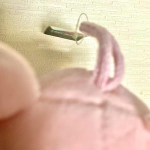 May include: A soft, light pink plush toy featuring a small, curled tail made of the same fuzzy fabric. A thin, white plastic fastener loop is attached to the top of the toy, holding a small rectangular tag with visible green and white text.