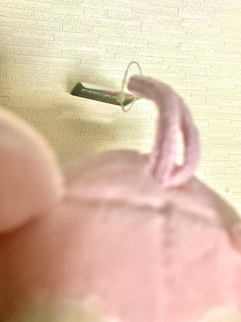 May include: A soft, light pink plush toy featuring a small, curled tail made of the same fuzzy fabric. A thin, white plastic fastener loop is attached to the top of the toy, holding a small rectangular tag with visible green and white text.