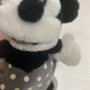 Op de afbeelding: Een Minnie Mouse knuffelpop met een zwart-wit gezicht, zwarte oren en een grijze jurk met witte stippen. De pop heeft witte handschoenen en een bruine strik op haar hoofd. Een klassiek Disney-personage.