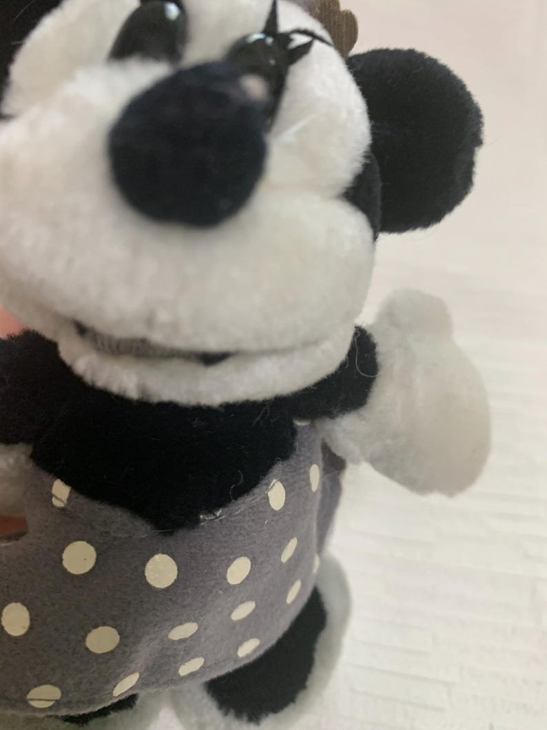 Op de afbeelding: Een Minnie Mouse knuffelpop met een zwart-wit gezicht, zwarte oren en een grijze jurk met witte stippen. De pop heeft witte handschoenen en een bruine strik op haar hoofd. Een klassiek Disney-personage.
