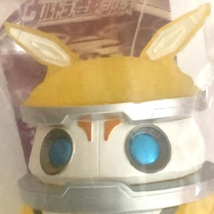 May include: A yellow and white plastic figure with two pointed ears, bright blue circular eyes, and silver metallic-colored trim around the head and neck. The figure is encased in clear packaging with Japanese text and the Bandai Namco logo visible.