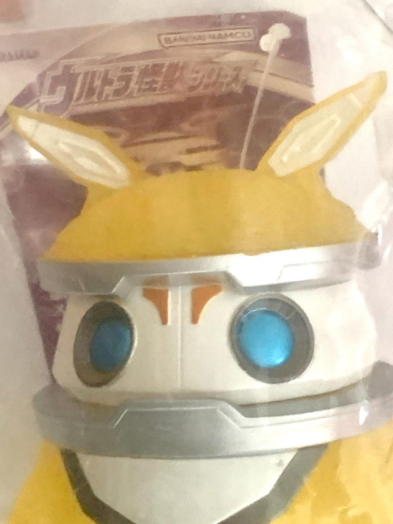 May include: A yellow and white plastic figure with two pointed ears, bright blue circular eyes, and silver metallic-colored trim around the head and neck. The figure is encased in clear packaging with Japanese text and the Bandai Namco logo visible.