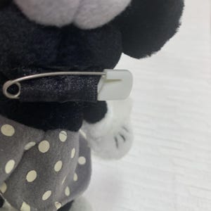 Op de afbeelding: Een Minnie Mouse knuffel met een zwarte strik vastgezet met een witte veiligheidsspeld. De pop heeft zwarte oren, een wit gezicht en een grijze rok met witte stippen. De achtergrond is een lichtgekleurd oppervlak.