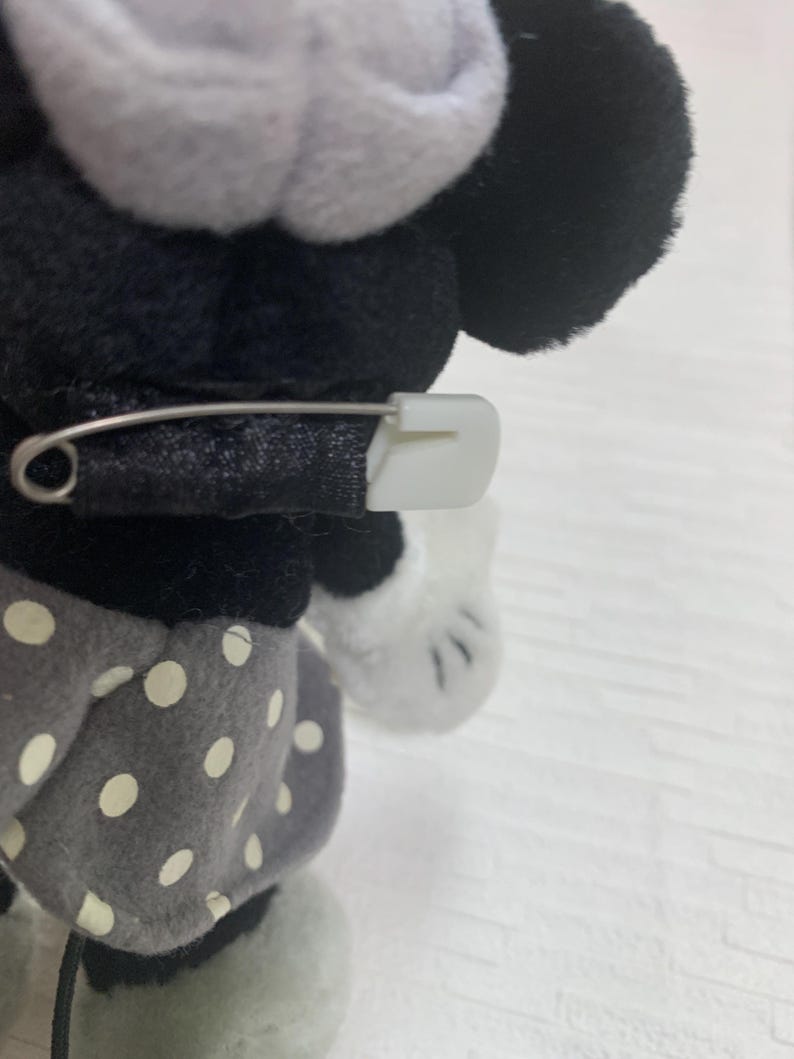 Op de afbeelding: Een Minnie Mouse knuffel met een zwarte strik vastgezet met een witte veiligheidsspeld. De pop heeft zwarte oren, een wit gezicht en een grijze rok met witte stippen. De achtergrond is een lichtgekleurd oppervlak.