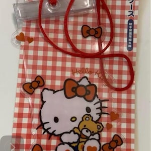 Pode incluir: Capa de telefone &agrave; prova d'&aacute;gua transparente com Hello Kitty segurando um ursinho de pel&uacute;cia, com la&ccedil;os e cora&ccedil;&otilde;es laranja em um fundo xadrez vermelho e branco. Inclui um cord&atilde;o vermelho. O texto na embalagem diz "Hello Kitty" e texto japon&ecirc;s.