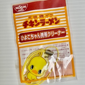 May include: A Nissin Chicken Ramen branded mobile phone cleaner charm featuring a yellow chick character. The item is enclosed in a clear plastic bag with orange and white striped packaging that includes Japanese text and the Nissin logo.
