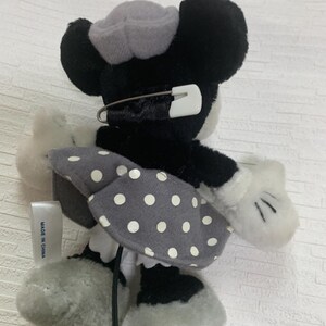 Op de afbeelding: Een Minnie Mouse pluche pop in zwart, wit en grijs. De pop heeft een grijze polkadot rok, een grijze hoed en witte handschoenen. Een veiligheidsspeld is aan het hoofd van de pop bevestigd. Het label luidt "MADE IN CHINA".