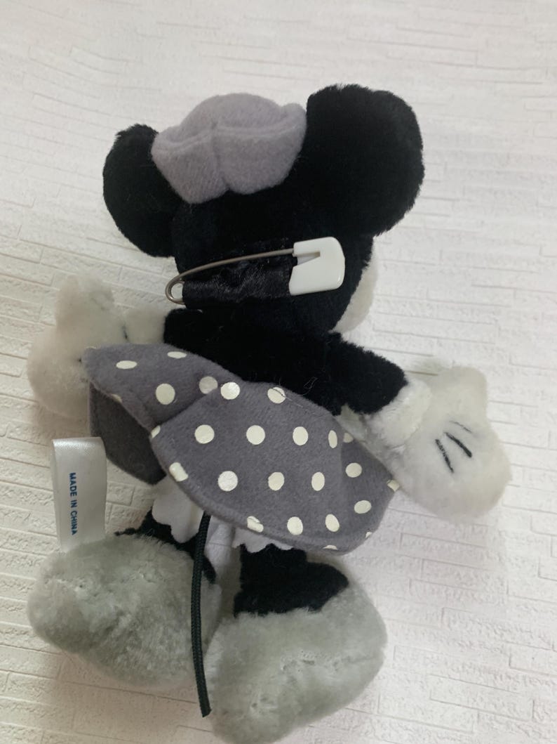 Op de afbeelding: Een Minnie Mouse pluche pop in zwart, wit en grijs. De pop heeft een grijze polkadot rok, een grijze hoed en witte handschoenen. Een veiligheidsspeld is aan het hoofd van de pop bevestigd. Het label luidt "MADE IN CHINA".